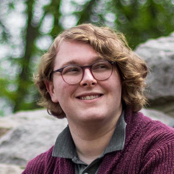 Sully wearing a collared shirt and cableknit sweater, smiling and looking away from the camera